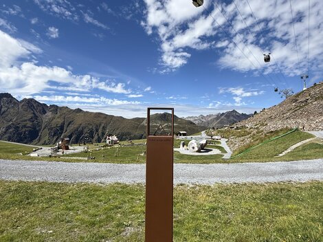 A mountain landscape with cable cars and a clear blue sky. In the foreground, there is a rectangular post in front of a green meadow.