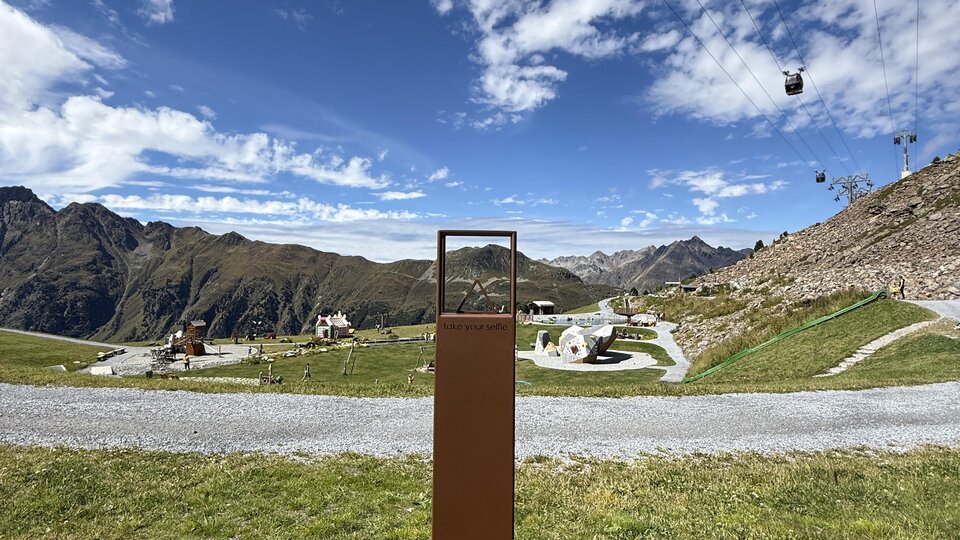 A mountain landscape with cable cars and a clear blue sky. In the foreground, there is a rectangular post in front of a green meadow.