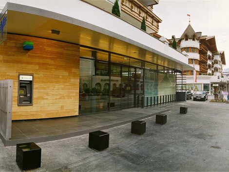 A modern building with large windows and wooden cladding. In the background, traditional architecture and mountains are visible.