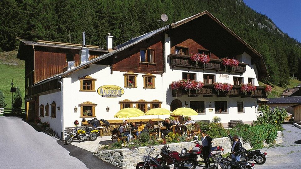 A cozy inn in the mountains with blooming balconies and sun umbrellas. Several motorcycles are parked in front of the building.