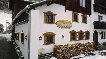 A traditional inn with a white facade and beautiful wood details. In the foreground, you can see a woodpile and a wintry landscape in the background.