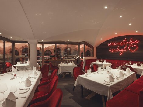 A stylish, warmly lit wine cellar with elegantly set tables, red velvet armchairs, and a glowing neon sign "Weinstube Ischgl" creates a cozy-exclusive atmosphere. | © Hannes Niederkofler