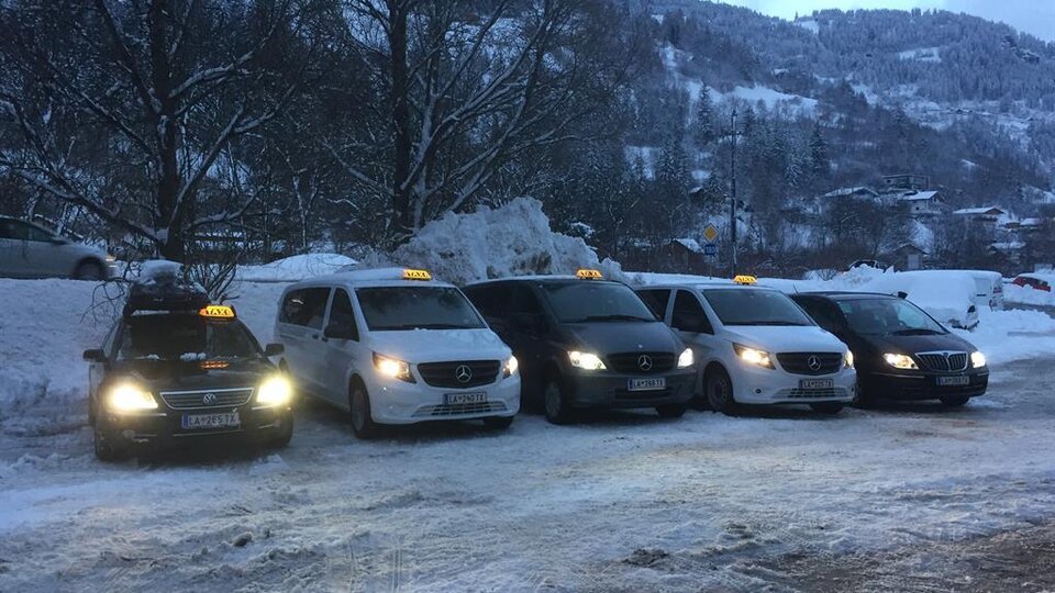 Fünf Taxis stehen in einer schneebedeckten Umgebung. Die Fahrzeuge sind in verschiedenen Farben und warten auf Passagiere.