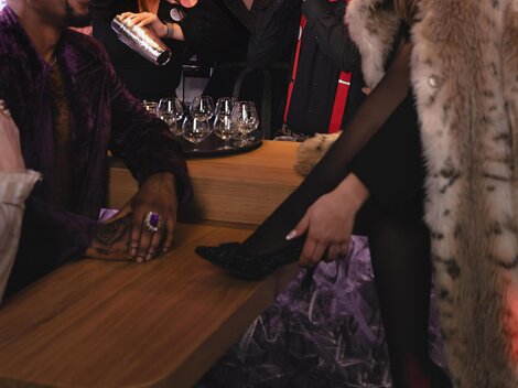 A vibrant bar scene with cheerful people mixing cocktails and an elegant woman in the foreground, sitting relaxed. The atmosphere is glamorous and modern, illuminated by colorful lights.