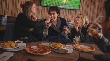 Eine Gruppe von Freunden sitzt in einem Restaurant und genießt Essen und Getränke. Im Hintergrund läuft ein Fußballspiel im Fernsehen.