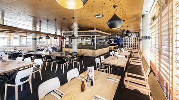A modern restaurant with light wood interiors and stylish lamps. The tables are neatly set and provide space for guests.