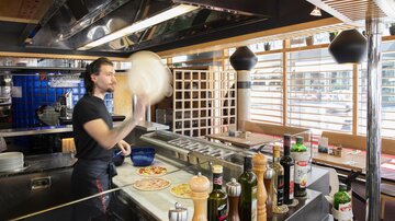 A chef in a modern pizzeria tosses pizza dough into the air. Various spices and ingredients are visible on the countertop.