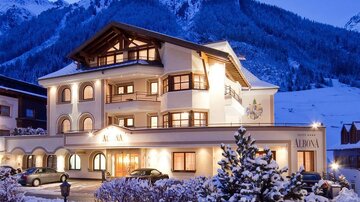 A charming hotel in the snow with a picturesque mountain panorama in the background. The facade is brightly lit and radiates an inviting atmosphere.