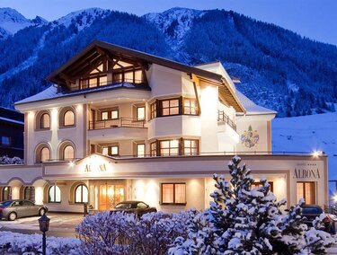 A charming hotel in the snow with a picturesque mountain panorama in the background. The facade is brightly lit and radiates an inviting atmosphere.