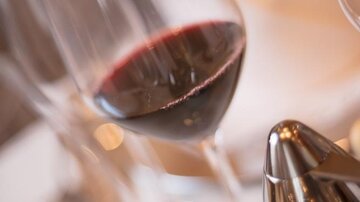 An elegant wine glass with red wine is standing on a table. In the background, additional glasses and cutlery are visible.