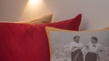 A cozy bed with red pillows and a decorative pillow featuring a black-and-white photo of two women skiing. A wall lamp provides soft light.