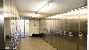 A narrow corridor with metal lockers on both sides. At the end of the corridor, there is a door that leads outside.