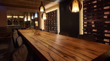 A stylish restaurant with a long wooden table and modern pendant lights. Numerous wine bottles are visible on the wall, creating a cozy atmosphere.