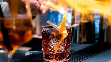 A cocktail is creatively prepared while fire burns above. In the background, other drinks are visible.