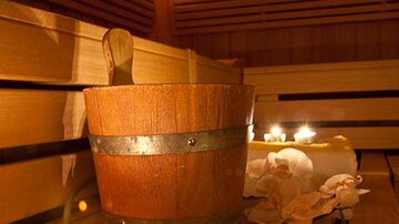 A wooden basket stands in a sauna, surrounded by wooden benches and candlelight. Relaxed atmosphere with orchid blooms for the feel-good experience.