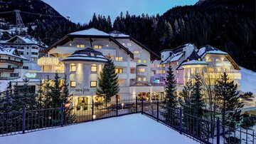 Ein elegantes Hotel in den Alpen, umgeben von schneebedeckten Bäumen. Die Fassade leuchtet sanft im Abendlicht.
