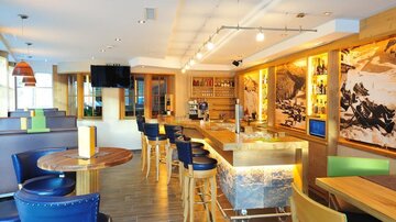 Ein moderner Innenraum mit Holzverkleidung und stilvollen Möbeln. Die Bar ist gut ausgestattet und der Raum hat große Fenster mit viel Tageslicht.