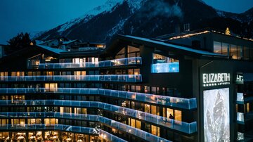Ein modernes Hotel mit beleuchteten Balkonen, umgeben von majestätischen Bergen. Der Abendhimmel verleiht der Szene eine ruhige Atmosphäre.