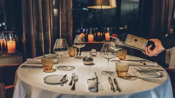Ein elegant gedeckter Tisch in einem Restaurant mit Kerzenlicht. Ein Kellner gießt Wein in ein Glas. | © elizabeth.at
