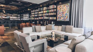 Eine moderne Lounge mit gemütlichen Sitzgelegenheiten und einer Bücherwand. Ein eleganter Tisch und farbenfrohe Kunstwerke schaffen eine einladende Atmosphäre. | © elizabeth.at