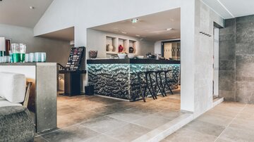 Eine moderne Bar mit stilvollem Design und einer hellen, einladenden Atmosphäre. Der Bereich ist mit eleganten Stühlen und einer schlichten Theke ausgestattet. | © elizabeth.at