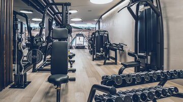 Ein modernes Fitnessstudio mit verschiedenen Trainingsgeräten. Im Vordergrund sind eine Hantelablage und eine Bank zu sehen. | © elizabeth.at