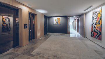 Ein moderner Flur mit Holzverkleidung und großen Bildern an den Wänden. Auf der linken Seite befinden sich Aufzüge, während der Raum hell und einladend wirkt. | © elizabeth.at