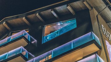 Ein modernes Gebäude mit beleuchteten Balkonen und einem markanten Fenster. Die Architektur wirkt stilvoll und einladend bei Nacht. | © elizabeth.at