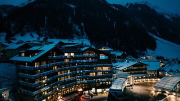 Ein modernes Hotel in den Bergen, umgeben von Schnee. Die Szenerie ist nachts beleuchtet und vermittelt eine gemütliche Atmosphäre.