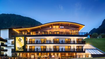 A modern hotel building with a illuminated roof and large balconies. The surroundings are characterized by hills and a clear evening atmosphere.