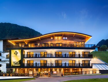 A modern hotel building with a illuminated roof and large balconies. The surroundings are characterized by hills and a clear evening atmosphere.