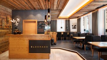 A modern restaurant with warm wooden decor and a stylish reception desk. Tables and chairs are comfortably arranged to create an inviting atmosphere.