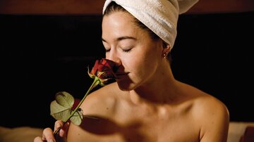 A woman with a towel on her head smells a red rose. She appears relaxed and content.