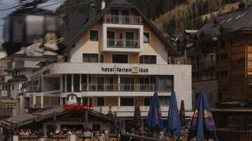 A modern hotel building in the mountains with terraces and a cable car connection. In the foreground, restaurant tables and sunshades can be seen.