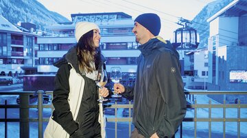 A couple stands outside holding glasses with drinks in hand. In the background, snow-covered mountains and a cable car can be seen.