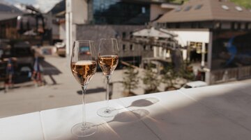 Two glasses of rosé sparkling wine are placed on a table, overlooking the mountainous landscape and buildings in the background. The scene conveys a sense of relaxation and enjoyment.