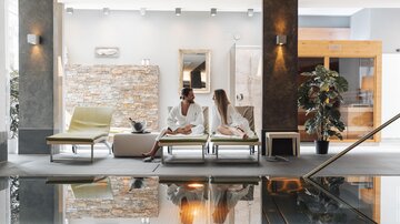 Ein eleganter Wellnessbereich mit Liegen und einem Wasserbecken. Zwei Frauen in Bademänteln entspannen und genießen die harmonische Atmosphäre.