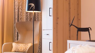 Ein eleganter Sessel steht neben einer Stehlampe in einem modernen Raum. Im Hintergrund sind Holzverkleidungen und ein Dekorationsobjekt sichtbar.