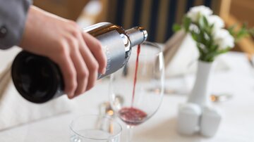 Ein elegantes Restaurant-Setting mit einem Glaskelch, in den gerade Wein eingeschenkt wird. Auf dem Tisch steht eine Vase mit frischen Blumen.