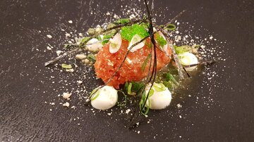 Ein kunstvoll angerichtetes Gericht mit einer Hauptzutat in der Mitte, umgeben von grünen Kräutern und kleinen weißen Saucenklecksen. Die Präsentation erfolgt auf einem schwarzen Teller, was den Farben des Essens Kontrast verleiht.