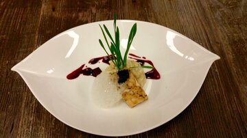 Ein kunstvoll angerichtetes Gericht auf einem weißen Teller. Es enthält Fisch, Reis, Kräuter und eine fruchtige Sauce.