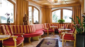 A cozy seating area with bright curtains and red-gold furnishings. Large windows let in plenty of daylight, creating an inviting atmosphere.