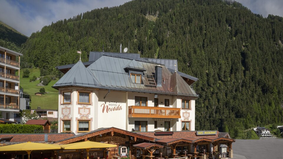 A charming hotel with a large balcony and an inviting terrace. In the background, green mountains and a cloudy sky can be seen.