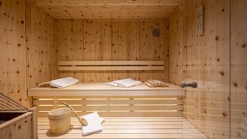 An elegant sauna made of light wood with a simple wooden bench. Towels and a sauna bucket are ready for the next wellness visit.