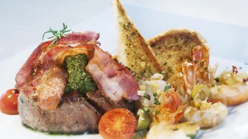 A plate with a juicy steak, topped with bacon and pesto, as well as shrimp and toasted bread. Cherry tomatoes are arranged alongside.