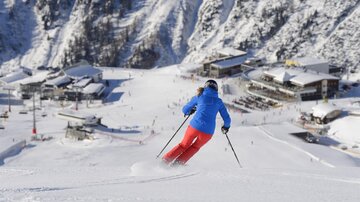 A person is skiing on a snow-covered slope. In the background, the mountains and a mountain hut can be seen.