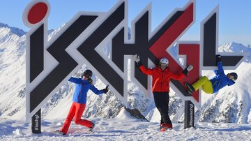 Three skiers are happily posing in front of a large sign that reads "Ischgl". In the background, snow-covered mountains and a blue sky can be seen.