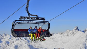 A cable car glides over snow-covered mountains. Three skiers are sitting relaxed in the gondola.