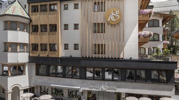 A modern hotel building with wooden cladding in a picturesque village. In the foreground, there are street cafes and floral arrangements.