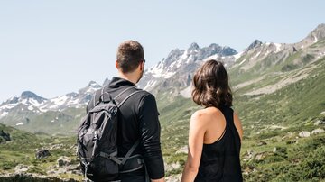 A couple looks at an impressive mountain landscape with snow-covered peaks. It is a sunny day and nature is lush and green.